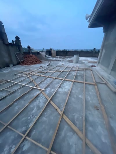 The image displays an elevated concrete platform or rooftop in Cutneuheun, Indonesia, under an overcast, blue-grey sky. A large grid of light-colored wooden battens lies directly on the textured, grey concrete surface, suggesting ongoing construction or groundwork. To the left, a mound of red bricks is situated beside a partially constructed concrete wall featuring ornamental, possibly finial-topped, structures. A rectangular frame leans against this wall. In the midground, a single white cylindrical container stands on the concrete. On the far right, a segment of a concrete structure with an overhang is visible, from which two white horn loudspeakers are mounted. The background reveals a low, dark-colored wall with a decorative, possibly crenellated, upper edge, behind which a flat, undeveloped landscape extends to the horizon under the same dim sky. No individuals are present in the scene.