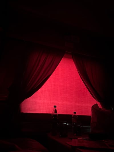This is a dimly lit indoor scene illuminated by a strong red light source, possibly a theatrical or decorative light. The image appears to be taken in Teykovo, Russia, though no specific landmarks are visible. The primary focus is a window covered by heavy, dark red curtains that are drawn back, revealing a bright red fabric behind them. This red fabric, likely a backdrop or another set of curtains, creates a stark, saturated color field.

In the foreground, on a surface that is mostly in shadow, are several objects. Two plastic bottles are visible, one on the left and one to the right of center. Both appear to contain liquid. Between them, and slightly in front, are what seem to be small, translucent plastic cups or containers. A white plastic bag is partially visible on the far right. The overall impression is one of a staged or prepared environment, possibly for an event or performance, given the dramatic lighting. No people are directly visible, though the presence of the objects suggests human activity. The environment is enclosed and dark, with the red light dominating the visual space.