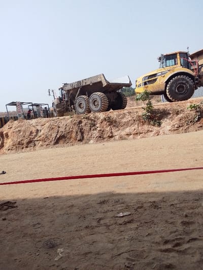 A construction site reveals two heavy-duty dump trucks parked atop a dirt mound. The first is a dark, well-used model with large tires. Adjacent to it is a bright yellow Volvo dump truck. In the background, a small structure houses a few individuals. A horizontal red line bisects the foreground, marking a boundary on the dirt ground.