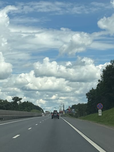 A black SUV is driving on a highway towards the horizon. The road is asphalt with white lane markings. There are trees and green grass on the right side of the road. A red and blue circular sign with a red cross is visible on the right side of the road, accompanied by a white sign with an arrow pointing upwards and the text "500 m". The sky is blue with white, puffy clouds. There are other cars and traffic lights visible in the distance. The scene appears to be in a rural or suburban area outside of the city of Leshkovo, Russia.