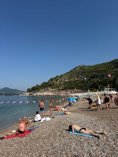 This media file depicts a sunny day at Kamenovo beach in Montenegro. The scene is a crowded pebble beach with many people sunbathing and relaxing. The water is calm and clear, with a few boats visible in the distance. A steep, tree-covered hill rises behind the beach, and a small town or resort can be seen nestled along the coastline further down.
The people are dressed in swimwear, and some are lying on towels or sun loungers. A few individuals are partially in the water. The atmosphere appears relaxed and leisurely, typical of a summer beach day. The time of day is likely mid-afternoon, given the bright sunlight and the shadows cast by the people.
Visible objects include beach umbrellas, towels in various colors, and a demarcation line with buoys in the water. There are several white structures that appear to be part of a beach setup or facilities. Some red and white signs are visible on the hillside, but the text is too small to read.
The overall impression is one of a vibrant and popular tourist destination, enjoying warm weather and a beautiful coastal landscape.