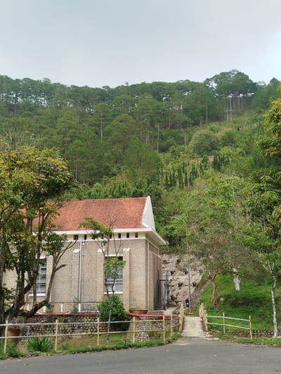 This image presents a tranquil, natural scene dominated by a building nestled against a densely forested hillside under an overcast sky.
The main subject is a two-story building with light-colored, possibly stone or brick, textured walls and a prominent terracotta-tiled hip roof. The upper part of the walls below the eaves is white, featuring small rectangular vents. Multiple windows with white frames are visible on the building's side, some appearing to be open louvre-style. A fence, made of light-colored wood or bamboo, runs along the foreground, separating a paved road from the building's immediate property.
To the right of the building, a narrow concrete path leads uphill, crossing a small, low-arched bridge with railings similar to the main fence. This path disappears into the thick vegetation covering the hillside. A set of black metal stairs is visible on the left side of the building, ascending along its wall. The hillside behind the building is steep and heavily wooded, primarily with tall, slender pine trees that extend to the horizon, creating a lush green backdrop. Several streetlights are intermittently visible among the trees on the slope.
The time of day appears to be daytime, likely a cloudy or overcast day given the diffused light and light grey sky. There are no people visible, and no specific activity is taking place, contributing to a serene and untouched atmosphere. The setting suggests a remote or natural location, possibly in a mountainous or hilly region. No visible text is present in the image.