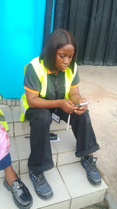 A woman wearing a bright yellow safety vest over a grey and orange polo shirt sits on concrete steps outdoors. She has dark, shoulder-length hair and is looking down at her silver smartphone, which she holds in her hands. She wears black pants that are cuffed at the ankles and dark athletic shoes. A beaded white bracelet hangs from her right wrist. A lanyard with identification cards is around her neck. To her left, another person's feet are visible, wearing black, shiny, loafer-style shoes. A black electronic device sits on the step below the woman's feet. The background features a bright blue wall and a dark, corrugated structure. The lighting suggests it is daytime, possibly overcast or the scene is in shade. The overall impression is one of a brief pause or break during a workday, with the woman engaged with her phone. Visible text on the lanyard includes "SAHCOL".