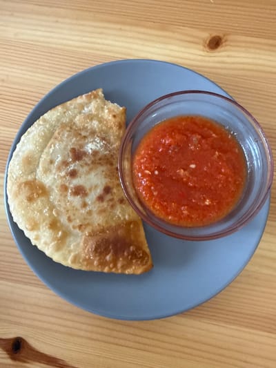 A top-down, close-up shot shows a gray plate on a light brown wooden table. On the plate, there is a small, clear glass bowl filled with a red sauce that appears to be made of pureed tomatoes and possibly spices or herbs. Next to the bowl, a golden-brown, fried pastry is placed. The pastry is folded and appears to be slightly crispy with some bubbly textures on its surface. The setting is casual, with the food arranged on a simple plate, suggesting a meal or snack. The lighting appears to be natural, possibly from a window, creating soft shadows and highlighting the textures of the food and the wood grain. No people are visible in the image. The context provided indicates the location is Zielona Góra, Poland.