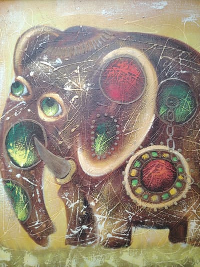 This is a close-up, horizontal, eye-level shot of an abstract painting on canvas featuring an elephant. The elephant is rendered in shades of brown, with several circular inlays on its body. These inlays contain various colors and textures. Some are a deep red, some a vibrant green with yellow accents, and one features a metallic chain link. The elephant's eyes are a striking emerald green. A single tusk protrudes from its face, and there are decorative, metallic-like studs along its forehead and body. The background is a soft yellow with abstract lines and splatters of white and gold paint overlaying the entire composition, creating a textured and somewhat chaotic effect. The painting appears to be an oil or acrylic on canvas. There is no visible text.