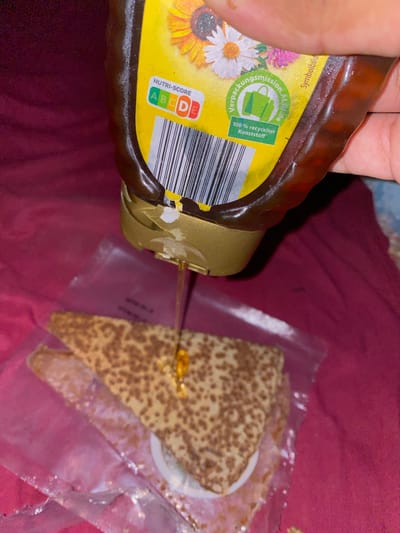 A close-up shot shows honey being drizzled from a yellow plastic bottle with a brown cap onto a slice of a baked good, possibly a cake, resting in a clear plastic bag. The cake appears to have a speckled brown pattern on its surface and is triangular in shape. The honey bottle features a Nutri-Score label with a "D" in red, indicating a less healthy nutritional profile. There are also labels with text in German, including "Verpackungsmission, Mitglied" and "100% recycelter Kunststoff," indicating that the packaging is part of a mission and made of 100% recycled plastic. The background is a dark red, textured surface, possibly fabric. The scene is likely taking place indoors, as suggested by the artificial lighting and the close proximity of the objects. There is no indication of a specific location in the city of Baraki, Algeria.