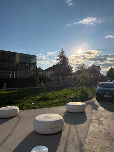 The image captures a scene in Ljubljana, Slovenia, featuring a modern building with a glass facade, a patch of green lawn, and a row of white, cylindrical bollards along a pedestrian path. A car is parked on a paved area to the right, partially obscuring buildings and trees in the background. The sky is bright blue with scattered white clouds and the sun is visible, casting long shadows on the pavement. The scene appears to be an urban landscape on a sunny day.