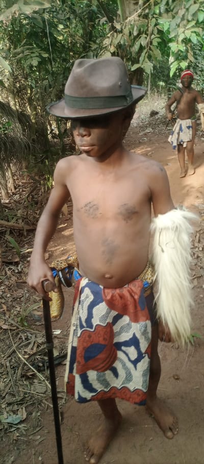 A child stands in a forest wearing a brown fedora, dark face paint, and tribal attire. The child has smudges of dark color on the chest and a white, fur-like arm covering. A patterned fabric wraps around the waist like a skirt. The child grips a dark walking stick. Another person in similar attire stands partially visible in the background, further back in the forest.