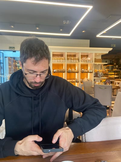 A man with dark hair and a beard is seated at a wooden table, wearing glasses and a black hoodie. He is holding a black smartphone in his hands and appears to be interacting with it. The man is sitting on a light-colored chair with decorative accents.
Behind him is a shelving unit filled with packaged food products, suggesting the location is likely a bakery or cafe. Through the glass in the background, a storefront entrance with Portuguese text ("SAIDA") can be seen. Geometric fluorescent lights are visible on the ceiling.
The image was taken indoors, during the daytime. Based on the environment, the location may be in Nova Petrópolis, Brazil, where Portuguese is the official language and bakery-style establishments are common.