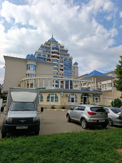 The image depicts a street scene in Goryachiy Klyuch, Russia, under a cloudy sky. The focal point is a modern, multi-story building with a distinctive pyramidal design and blue accents on its roof and windows. The building appears to house several businesses, with visible signage indicating "ФМOTECT МЕДИЦИНСКАЯ ЛАБОРАТОРИЯ" (FMOTECT MEDICAL LABORATORY) and "ЦВЕТЫ" (FLOWERS), along with "Студия ПАРИКМАХЕРСКАЯ" (Studio HAIRDRESSER) and "КНАЛЕРСКАЯ" (KNALERSKAYA), likely a salon.
In the foreground, a silver van with the license plate "H290BX 193" is parked. To the right of the van, a silver Kia Sportage SUV is parked, displaying the license plate "Y163PM 123". Another silver car is partially visible to the right of the Kia. The area in front of the building has a paved surface with yellow bollards and is bordered by a strip of green grass. The architecture of the building suggests a relatively modern construction, and the overall atmosphere appears calm, with no people visible in the immediate vicinity. The presence of shops suggests it is likely daytime, and the cloudy sky indicates the weather is overcast.