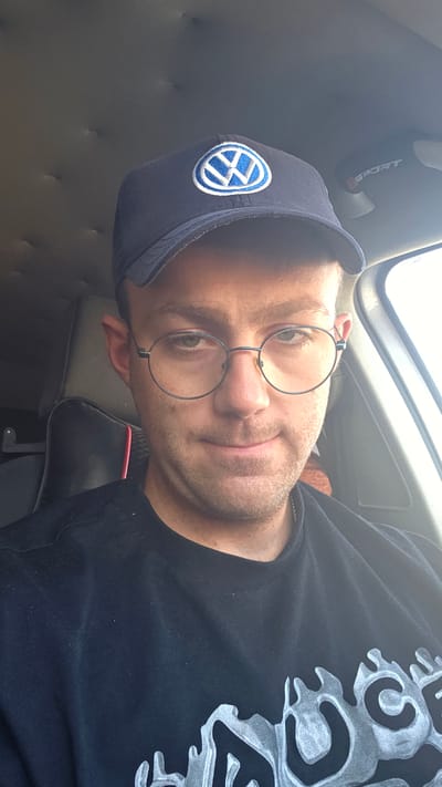 A man is seated in the driver's seat of a car, looking directly at the camera. He is wearing a dark blue baseball cap with a blue and white Volkswagen logo. He also has on round, wire-rimmed glasses and a black t-shirt with a graphic design. The background shows the interior of the car, including the car's ceiling, a headrest, and part of the windshield. The lighting suggests it is daytime. The image was taken in Bania, Bulgaria.