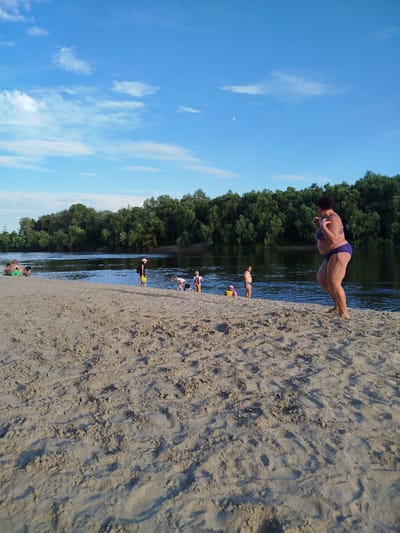 A woman in a purple bikini stands on a sandy riverbank, her left hand on her hip and her right arm bent upwards. She is facing left, looking out towards the river. Behind her, a dense line of trees forms the opposite bank of the river, under a clear blue sky with some scattered white clouds and a sliver of the moon visible. Several children are scattered across the sandy foreground and along the water's edge, some playing and others walking. A man in dark shorts stands in the shallow water on the left, and a few people are seated on the sand near the water. The scene appears to be a public beach in Chernihiv, Ukraine, on a warm day.