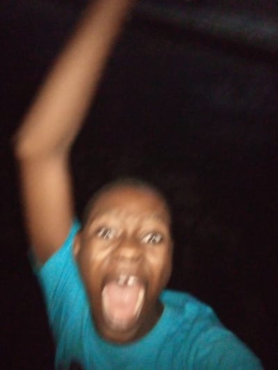 This is a blurry, close-up, waist-up photograph taken in low light conditions. The subject is a young person, possibly a child, wearing a bright teal t-shirt. The subject's mouth is open wide in what appears to be a scream or a shout, with their tongue visible. Their eyes are open and directed towards the camera, and their left arm is raised and blurred, suggesting movement. The background is uniformly dark and out of focus. The image quality is poor due to motion blur and low light. There is no discernible indication of the city of Shangisha, Nigeria, within the image itself.