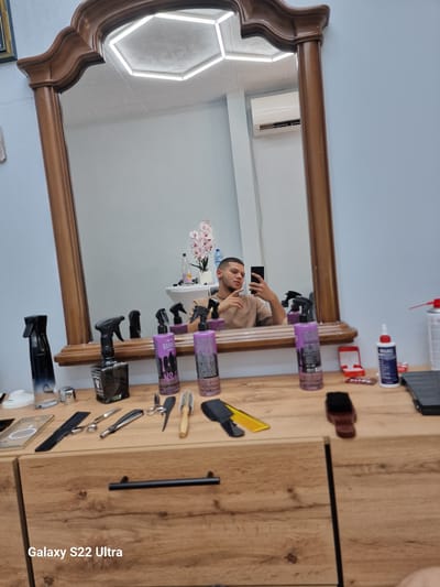 A young man with short dark hair and a light brown t-shirt takes a selfie in a barber shop. He is holding a black smartphone in his right hand, with his left hand resting on his chin. The man is reflected in a large ornate mirror that dominates the frame. In the reflection, on a shelf behind the man, is a vase of pink orchids and some bottles of liquid, including a bottle of water.
In the foreground, on a wooden dresser, are various barbering tools and products, including spray bottles, scissors, a razor, and combs. The dresser has two drawers with black handles. The wall behind the mirror is a light blue. A white air conditioning unit is visible in the reflection on the back wall. The text "Galaxy S22 Ultra" is overlaid in the bottom left corner of the image. The scene appears to be a barber shop in Vidin, Bulgaria.