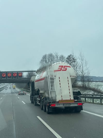 The image displays a multi-lane German autobahn on an overcast day, likely during winter or late autumn, near Irschenberg. The road surface is wet, and patches of snow are visible on the ground beside the highway. In the right foreground, a large articulated truck with a dark cab is traveling, pulling a white silo-trailer. The trailer bears the red logo "BS" and the text "BETON-SCHMITT". Dark exhaust fumes are visible emanating from the truck's exhaust stack. A metal ladder is affixed to the side of the silo-trailer. The rear of the trailer displays a license plate "MZ BP 555" and illuminated red taillights. To the left of the truck, a white passenger car, possibly an older model, is seen towing a small, single-axle trailer. Overhead, a gantry spans the highway, equipped with four illuminated digital signs. Three of these signs display a speed limit of "80" km/h, while the fourth indicates a "no overtaking for trucks" restriction with a red circle and truck icon. The right side of the highway is bordered by a metal guardrail, beyond which bare deciduous trees, some evergreens, and snow-dusted fields are visible. Additional vehicles are present further down the highway in the distance. No individuals are clearly identifiable.