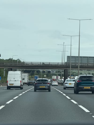 The image depicts a view from a vehicle traveling on a multi-lane motorway, likely the M25 in the city of Hertsmere, United Kingdom, as indicated by the presence of UK license plates and road markings consistent with British motorways. The sky is overcast and a light grey. The motorway appears to be experiencing moderate traffic with several cars, vans, and trucks visible. In the foreground, a white van is on the left lane, followed by a dark colored hatchback in the middle lane. To the right, a dark teal SUV is in the right lane. Further ahead, an overpass with a concrete barrier and railings stretches across the motorway. Tall lamp posts with multiple fixtures are positioned along the side of the road. Signs and other vehicles are visible in the distance, suggesting ongoing traffic flow. The road surface is asphalt with clear white lane markings. The environment appears to be rural or semi-rural, with trees visible beyond the overpass.