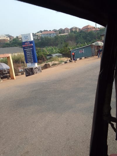The scene depicts a daylight street view in Nkwelle Ezunaka, Nigeria, captured from within a vehicle. The asphalt road surface occupies the foreground and mid-ground. To the left, a light-colored archway stands next to a parked motorcycle under an awning. Centered mid-left, a prominent blue and white vertical sign displays "THIS IS RIVERBANK ESTATE" along with a list of addresses. To the sign's right, a dirt shoulder with black and white striped curbing borders the asphalt. On this shoulder, a small, blue corrugated metal shack is visible, along with another structure partially obscured by an umbrella. One individual, wearing dark shorts and a shirt, walks rightward on the dirt shoulder, carrying an orange container on their left shoulder. Two additional figures are indistinctly visible near the blue shack. The background features a densely vegetated hillside dotted with numerous multi-story residential buildings exhibiting various roof colors (e.g., red, brown, grey). Dark structural components of the viewing vehicle frame the top and right edges of the image.