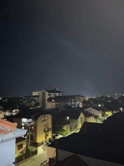 This is a nighttime aerial photograph of a residential area in the city of Cacak, Serbia. The image captures a densely populated neighborhood with a mix of multi-story apartment buildings and smaller houses. The dark night sky is punctuated by a few faint lights in the distance, possibly from streetlights or distant buildings. The buildings are illuminated by interior lights, creating a warm glow from windows. Some structures have visible balconies and rooftops, with the characteristic red tile roofs common in the region. The foreground features the edge of a building or balcony, with a white gutter and a section of reddish-orange roof tiles. The overall scene depicts a quiet, urban evening with signs of life visible through the illuminated windows of the homes.