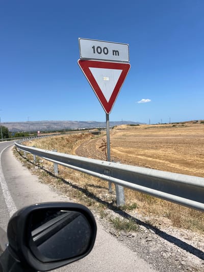 The image captures a roadside scene under a clear blue sky in the vicinity of Manfredonia, Italy. Dominating the foreground is a road sign displaying "100 m" above a red-bordered yield sign. A metal guardrail runs alongside the road, separating it from a dry, grassy field. In the lower left corner, a portion of a car's side mirror is visible. The landscape beyond the field features rolling hills with some scattered vegetation and power lines. The overall environment suggests a rural or semi-rural setting.