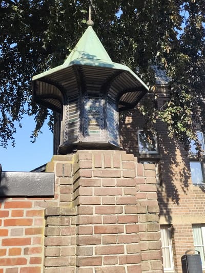 The image shows a decorative structure atop a brick pillar, located at the Kerkstraat in Schaijk, Netherlands. The structure features a green, octagonal roof and a lantern-like enclosure with patterned glass panels. The pillar is constructed from red and grey bricks, with a dark, rectangular element attached to its left side. Behind the pillar, a brick building with windows is visible, partially obscured by the leaves of a large tree. The sky is clear and blue, indicating it is daytime. There are no people or animals present in the image.