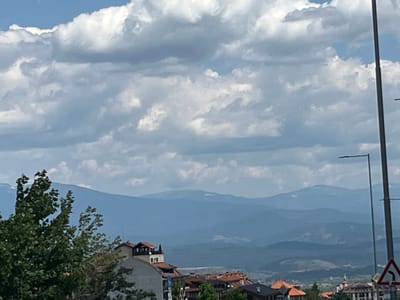 The image displays a panoramic view of Bansko, Bulgaria, under a sky filled with cumulus clouds. In the foreground, lush green trees are visible on the left side, with a cluster of residential buildings with orange and brown tiled roofs situated in the mid-ground. Some buildings have white facades, while others are a lighter grey. A tall, dark lamppost stands prominently on the right side of the frame, with a traffic sign indicating a sharp left turn. In the background, multiple layers of blue mountains recede into the distance, creating a sense of depth. The overall scene suggests a bright, possibly breezy day in a mountainous region.