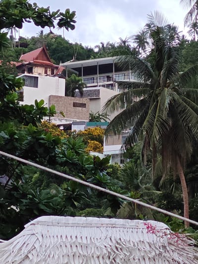 The image depicts a view of buildings nestled into a lush, tropical hillside in Baan Chaweng Noi, Thailand. In the foreground, a portion of a white, thatched roof is visible, with a thin rope extending diagonally across the frame. Behind this, dense green foliage and the fronds of a prominent palm tree dominate the mid-ground. Several modern buildings with white facades, balconies, and dark-tinted windows are interspersed among the greenery and on the slope. One building on the left features a distinctive red, tiered roof with ornate gables, suggesting a Thai architectural style. The background consists of more densely vegetated hills and a cloudy sky. The scene conveys a sense of tropical seclusion and resort living.