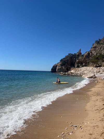 The image shows a beach scene at Cala del Moraig in El Poble Nou de Benitatxell/Benitachell, Spain. A rocky cliff rises on the left side of the beach. A person with a black cap and dark swimming trunks is standing in the water with a yellow surfboard. The water meets the sandy beach with foamy waves. The sky is clear and blue. The image is taken at an angle.