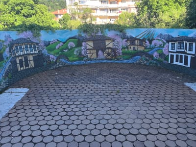 The image depicts a mural on a curved wall in the city of Troyan, Bulgaria. The mural features a picturesque landscape with traditional Bulgarian houses, a watermill, a stream, mountains, and blooming trees. The foreground is a paved area with circular grey paving stones. Behind the wall, lush green trees and apartment buildings are visible. The overall scene is bright and appears to be taken on a sunny day. There are no people or animals visible in the image.
