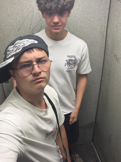 The image shows two young people, likely male, in a close-up, portrait-style shot. Both are wearing white t-shirts. The person in the foreground is wearing a black baseball cap with a flame-like design and round-rimmed glasses. The person in the background has curly brown hair and is looking slightly away from the camera. The setting appears to be indoors, possibly a locker room or a similarly utilitarian space, with textured grey walls. There are some faint markings on the wall in the background that are illegible. The image is oriented sideways. No specific activity is clearly depicted, and the lighting suggests it could be daytime, but no external weather cues are present. The overall mood is casual and candid.