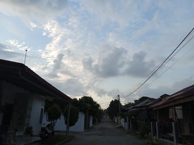 The image depicts a residential street in Buketteukuh, Indonesia, under a partly cloudy sky. On the left, a white house with a dark, sloped roof is visible, featuring a covered porch with a motorcycle parked on it. A street lamp stands near the edge of the property. On the right side of the street, several houses with red, sloped roofs are shown, some with open porches and fences. The road surface is paved and appears to be a narrow street winding into the distance, where a single red light, likely from a vehicle, is faintly visible. The surrounding environment includes mature trees with dense foliage lining the street and between the houses. Power lines are strung across the upper portion of the frame. The overall scene suggests a quiet, possibly early morning or late afternoon atmosphere in a suburban or rural Indonesian setting.