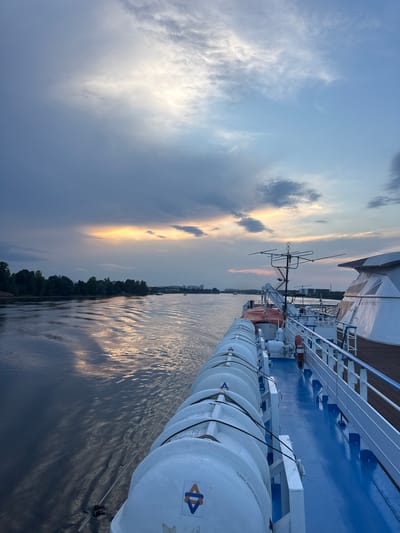The image captures a view from the deck of a ship at sunset, looking out over a wide river. The sky is dramatic with a mix of dark, heavy clouds and areas where the setting sun is breaking through, casting a warm, golden light. The water reflects the sky, creating shimmering patterns. On the right side of the image, the ship's deck is visible, painted bright blue, with a row of white life rafts neatly secured along the railing. The railing itself is white, with a wooden handrail. A life preserver hangs on a stanchion. In the distance, a tree-lined shore is visible, with a city skyline appearing on the horizon. The scene is serene and suggests travel on a calm waterway. The location is the city of Imeni Sverdlova, Russia, which is situated on the Neva River, a major waterway.