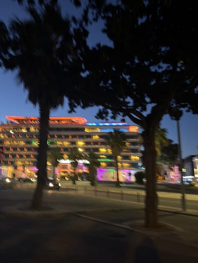 The image shows a blurred nighttime scene of a building with illuminated windows and colorful lights. A dark silhouette of a tree is in the foreground, partially obscuring the view of the building. The lights on the building are streaks of various colors, including orange, red, yellow, green, and purple, suggesting a vibrant urban environment at night. The sky is a dark blue, indicating dusk or nighttime. The reflection of the building and lights can be seen in what appears to be a body of water or a wet surface below, adding a symmetrical element to the composition. The overall impression is one of a lively city or resort area after dark. The motion blur suggests the photo was taken while moving, perhaps from a vehicle or on foot. There is no visible text in the image.