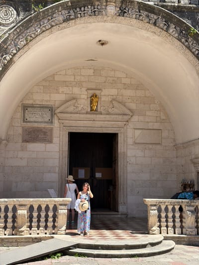 The image depicts the exterior of the Church of St. Luke in Kotor, Montenegro, on a sunny day. In the foreground, a stone ramp leads up to a checkered tiled area in front of the church entrance. A woman in a blue floral dress and a straw hat is holding another straw hat and looking at the camera. Behind her, to the left, another woman in a white dress and a straw hat stands near an information board. The facade of the church is made of stone blocks, with a large arched entrance. Above the entrance, there is an ornate stone carving and a golden statue. Two plaques are mounted on the wall to the left of the entrance. To the right of the entrance, a stone railing with decorative carvings lines the walkway. In the background, to the far right, a person is leaning over the railing near a display of small items.