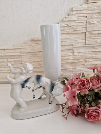 This is a close-up, eye-level, indoor photograph of decorative objects on a white surface. In the foreground, on the left, is a white porcelain figurine of a cherubic child interacting with a small, dark-and-white spotted animal, possibly a lamb. The child is in a playful pose, with arms outstretched, while the animal is on its hind legs, appearing to engage with the child. To the right of the figurine, there is a bouquet of pink artificial flowers. In the background, a tall, fluted white vase stands behind the figurine. The wall behind the objects has a textured, off-white stone veneer arranged in staggered horizontal rows. The overall impression is of a still life arrangement. The image does not contain any identifiable landmarks or details that would suggest a specific location within the city of Bremen, Germany.
