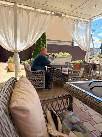 In Leningradskaya, Russia, a bald man wearing a dark teal short-sleeved t-shirt and dark jeans is seated at a woven patio table and chairs, facing away from the camera. He is holding his hand to his chin as if in thought. The setting appears to be an outdoor cafe, with white fabric draped from a pergola overhead. Other woven chairs are visible, one with an orange throw blanket draped over it. The ground is paved with tan and rose-colored rectangular tiles. In the background, a brown wooden fence and some greenery are visible, along with a building. The sky is blue with scattered white clouds. The lighting suggests it is daytime.