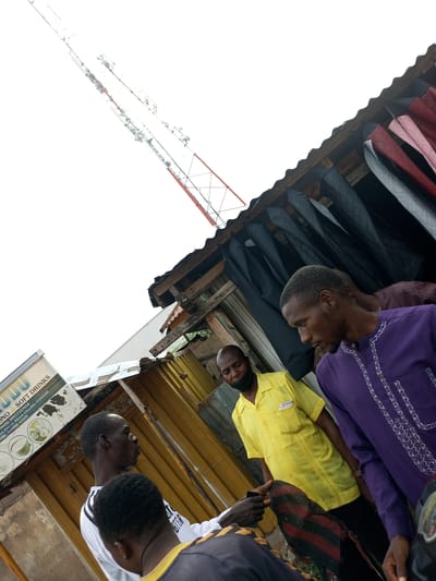 In Arawa, Nigeria, this image captures an outdoor scene with a telecommunications tower prominent in the background against a bright sky. In the foreground, several individuals are gathered near a makeshift structure that appears to be a market stall, with clothing items, possibly suits or jackets, displayed under a corrugated metal roof. One man in a purple garment is looking downwards, while another in a yellow shirt is facing the camera with a black face mask pulled down. A third man, wearing a white shirt with black stripes, is partially visible. The environment suggests a bustling, informal commercial area.