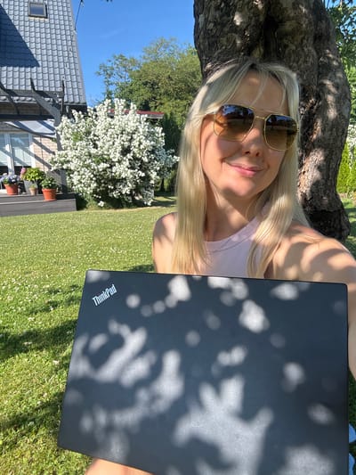 A woman with blonde hair and sunglasses is lying down outdoors, propped against a tree. She is holding a closed black laptop on her lap. The laptop has a white "ThinkPad" logo on it. She is wearing a light pink tank top. The background shows a lush green lawn with white wildflowers, a large bush with white flowers, trees, a house with a grey tiled roof, and a clear blue sky. The sunlight filters through the tree leaves, casting dappled shadows on the woman and the laptop. The scene suggests a pleasant, sunny day, likely in the late morning or early afternoon, given the bright sunlight and clear sky. The overall mood is relaxed and peaceful, with the woman enjoying a moment outdoors, possibly working or taking a break. The location is identified as Carnikava, Latvia.