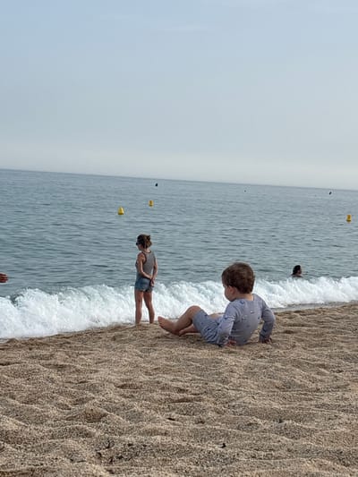 A child, seen from behind, is sitting on a sandy beach in Vilassar de Mar, Spain. The child is wearing a light blue pajamas with a pattern of ships and is leaning on their hands. In the background, waves crash on the shore. A woman wearing a striped tank top and denim shorts stands in the shallow water, facing away from the camera. In the distance, several yellow buoys are visible in the sea. The sky is overcast.