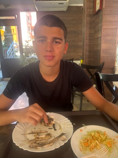 A young man is eating a meal at a restaurant. The man has short, dark hair and is wearing a black shirt. In front of him is a tray with two plates. One plate has leftover fish bones on it and the other plate has salad. The man is holding a piece of fish in his hand. Behind him, there is a window to the outside showing a ladder truck and text written in Cyrillic. The restaurant has wood paneling and a black chair. The text visible through the window, "СЛ. АХОВ ОЖЕ", suggests that the location is likely in Sandanski, Bulgaria, as that is where Slaviya Akhov OGE School is located.