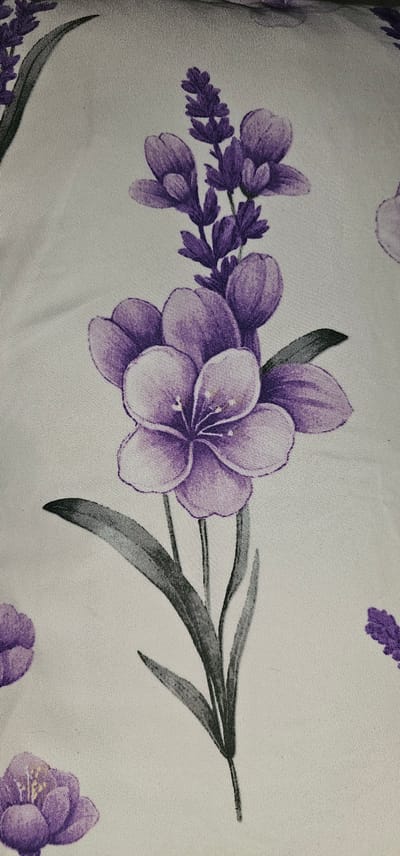 The image displays a fabric with a floral pattern. The primary subjects are stylized purple flowers, some resembling lavender sprigs and others appearing more like larger, open blossoms with delicate petals and stamen. These flowers are arranged in a decorative motif across a light-colored background, likely a textile like cotton or polyester, given the visible weave texture. The color palette consists of shades of purple for the flowers, with accents of green and grey in the leaves and stems. The pattern is repeated across the fabric, suggesting it could be part of a larger piece of material like a tablecloth, pillowcase, or garment. There are no people or specific scenes depicted, only the repeating floral design. The lighting appears even, with no strong shadows, typical of indoor photography or diffused natural light. No text is visible within the image. The location context of Makati, Philippines, does not directly influence the visual description of the fabric's pattern itself.