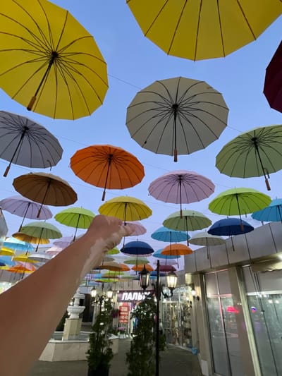 The image depicts an outdoor scene featuring numerous colorful umbrellas suspended in the air against a clear blue sky. In the foreground, a person's arm and hand are visible, reaching towards the umbrellas. The hand has patches of vitiligo. Below the umbrellas, a street or walkway is lined with buildings that appear to be shops or commercial establishments. A fountain is visible on the left side of the scene, and streetlights are present. Signs in Cyrillic script are visible on the buildings, one of which reads "ПАЛЬ". The setting appears to be a public space in a city.