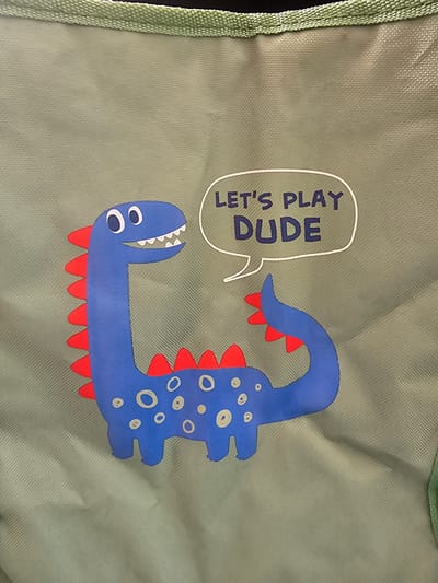 A close-up, eye-level, medium shot captures a stylized blue cartoon dinosaur with red spikes along its back and tail, set against a textured, light olive-green fabric background. The dinosaur has large, round white eyes with black pupils and a wide, toothy grin. Above its head, a white speech bubble contains the text "LET'S PLAY DUDE" in dark blue, block lettering. The fabric has a visible woven texture and a light green edge trim is visible at the top of the image. The scene is devoid of any people or discernible elements of the city of Holon, Israel.