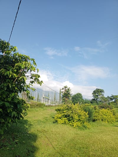 The image depicts a lush, green landscape under a blue sky with scattered clouds. In the foreground, a tree with dense green foliage is present on the left. The foreground transitions into a grassy field with a few bushes. In the mid-ground, there is a white fence and a sign that reads "MALANGKE BULANSE HOCKEY OLYMPIC CENTER." Mountains are visible in the background. Overhead, power lines bisect the sky. The specific location within Thalang, Laos, cannot be confirmed based on the visible details.