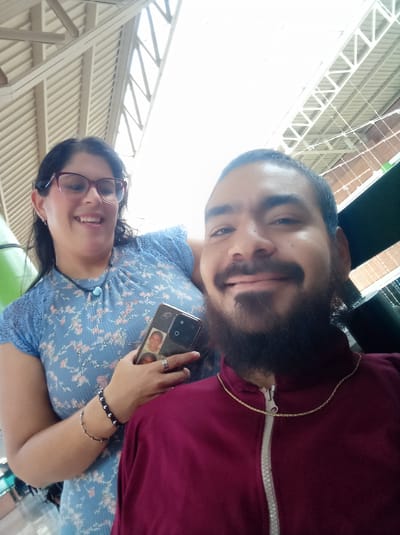 The image shows two people in what appears to be an indoor public space. The woman on the left wears a light blue floral dress and eyeglasses. She is holding a smartphone in her hand, which displays images of two young women. The man on the right wears a maroon jacket with a gold chain and has a beard. They are both smiling. The background features a ceiling with visible support beams and what seems to be the exterior of a building. Based on the location request, the scene is located in Parroquia Guatire, Venezuela, but no location specific indicators exist in the photo.
