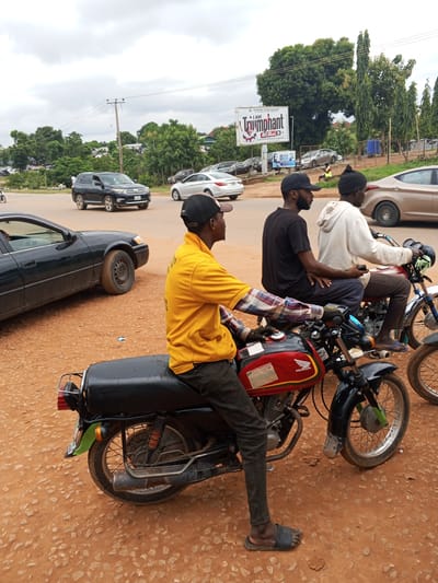 In Galadimawa Village, Nigeria, a medium shot captures an overcast day with three men on motorcycles at a street intersection. The primary focus is on a motorcycle in the foreground, ridden by a man in a bright yellow shirt and dark pants, wearing a black baseball cap. He is looking to his left, with his right arm extended, suggesting he might be signaling or adjusting something. Behind him, on a second motorcycle, two men are seated. The driver wears a black t-shirt and a black beanie, while the passenger behind him is in a white hooded sweatshirt. Their interaction is unclear from the image.
The environment consists of a dirt road with loose gravel and sand. In the background, several cars are visible, including a dark SUV and a beige sedan. Trees and foliage line the sides of the road, and a large billboard with the word "Triumphant" is prominently displayed. Power lines run overhead, indicating infrastructure in the area. The overall impression is a typical street scene in a Nigerian village.