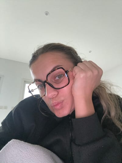 An indoor, close-up image taken in Veauchette, France, displays a female subject from the chest up. She wears a dark-colored, zippered top and large, square, tortoiseshell-patterned eyeglasses with gold accents at the temples. Her light brown hair is partially visible, and her eyes are directed forward. She rests the right side of her face on her right hand, with her fingers visible near her temple, while her lips are pursed in a puckered expression. A light-colored fabric is visible in the lower left foreground. The background consists of a plain, light-colored wall with a circular ceiling fixture and rectangular wall fixtures visible to the left.