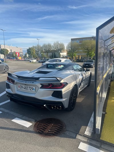 The image captures a silver Chevrolet Corvette C8 sports car parked in an outdoor lot on a clear, sunny day in Volketswil, Switzerland. The car is viewed from the rear-right, showcasing its sleek design, black wheels, and a subtle rear spoiler. A visible Swiss license plate on the car reads "ZH-482729", indicating its registration in the canton of Zurich.
The parking area features asphalt pavement with white parking lines and a circular metal manhole cover visible near the rear of the Corvette. A yellow-painted curb or barrier runs along one side of the lot, next to a transparent, shelter-like structure that lines the top of the frame.
In the background, beneath a bright blue sky with scattered wispy clouds, several commercial buildings are visible. One prominent building displays the word "amicasa" on its facade, partially obscured by green trees. Further in the distance, another building is identifiable by a red "KFC" sign, and inverted text, possibly "CHAJIAO", is also discernible. Other parked cars and streetlights are scattered throughout the background. The strong sunlight casts distinct shadows, suggesting the time of day is late morning or early afternoon. No people are visible in the scene.