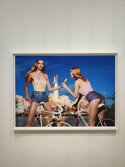 This is a photograph of two women on bicycles, in front of a launching space shuttle and a body of water. The woman on the left is wearing a yellow tank top with a picture of a crown and face on it, and blue sequined shorts. She has blonde hair and is looking at the camera. The woman on the right is wearing a pink tank top and blue sequined shorts. She is holding an orange and yellow popsicle to her mouth. Both women are wearing sheer stockings and are in a pose as if they are about to ride their bicycles. The space shuttle is in the background, appearing to be in the process of launching, with fire and smoke coming from its base. The sky is bright blue with some clouds. The body of water in the background suggests this scene is near the coast. The photograph is framed and hanging on a white wall, with a small label below it that reads "Weil Ranallly Razuco is Cocaine" and further text in a smaller font. This photograph is displayed in a museum in The Hague, Netherlands.
