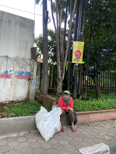 An adult female wearing a red shirt, green cap and a face mask sits on a sidewalk in front of a white concrete wall and a large tree, with a white plastic bag beside her. The scene is in Kecamatan Cimahi Selatan, Indonesia at latitude: -6.901, longitude: 107.563. She is looking directly at the camera. There is a yellow sign attached to a pole behind her with the text "Maxim" and "Hemar terus tanpa promo!" which translates to "Always save without promotions!" in Indonesian. The sidewalk is made of gray concrete blocks. The scene is illuminated by bright sunlight and appears to be in a residential area with a wooden fence and lush vegetation. The tree is a common species in Indonesia, and the wooden fence is a typical construction material in the region. The white concrete wall is likely part of a residential building or public structure. The scene is a glimpse into the everyday life of the people of Kecamatan Cimahi Selatan, Indonesia.