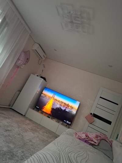 The image shows a bedroom with a TV, a refrigerator, a door, and a bed. The room is decorated in a modern style with light-colored walls and furniture. The TV is displaying a picture of a city at sunset. The bed is covered with a pink and white comforter. There is also a pink balloon with a cartoon character on it tied to the curtain. The scene is lit by natural light from a window and artificial light from a ceiling fixture and a table lamp. The temperature displayed on the TV screen is 19 degrees Celsius. The time on the TV is 20:47.